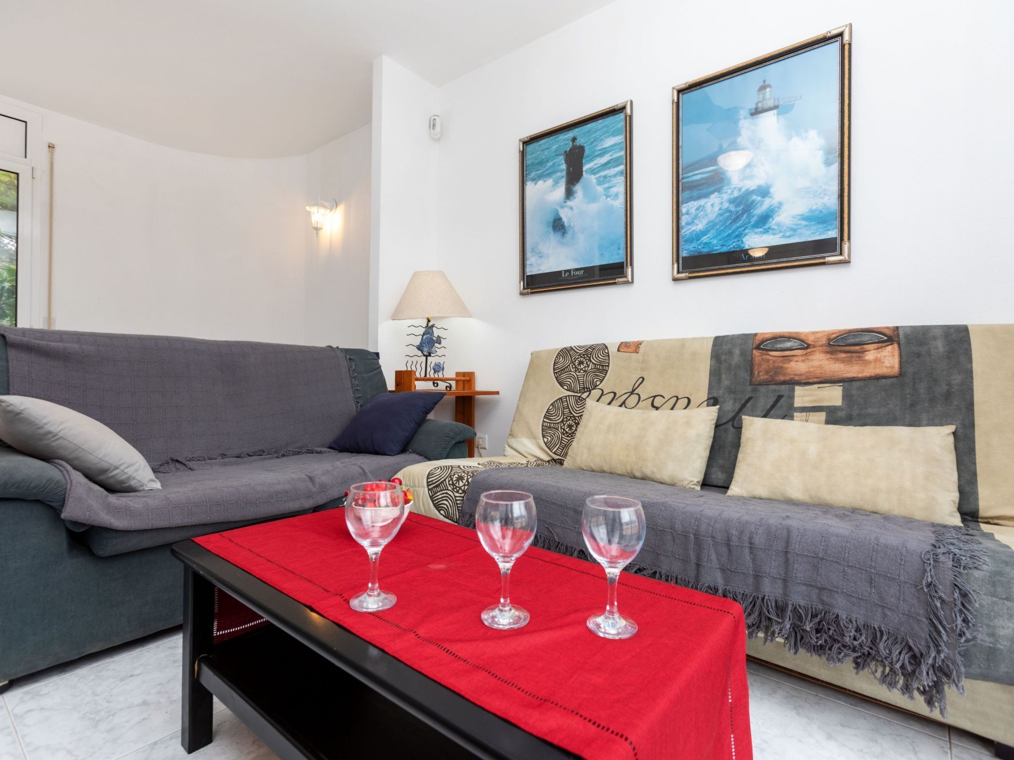 A living room with a grey sofa, a red table with three wine glasses, and two framed pictures on the wall.