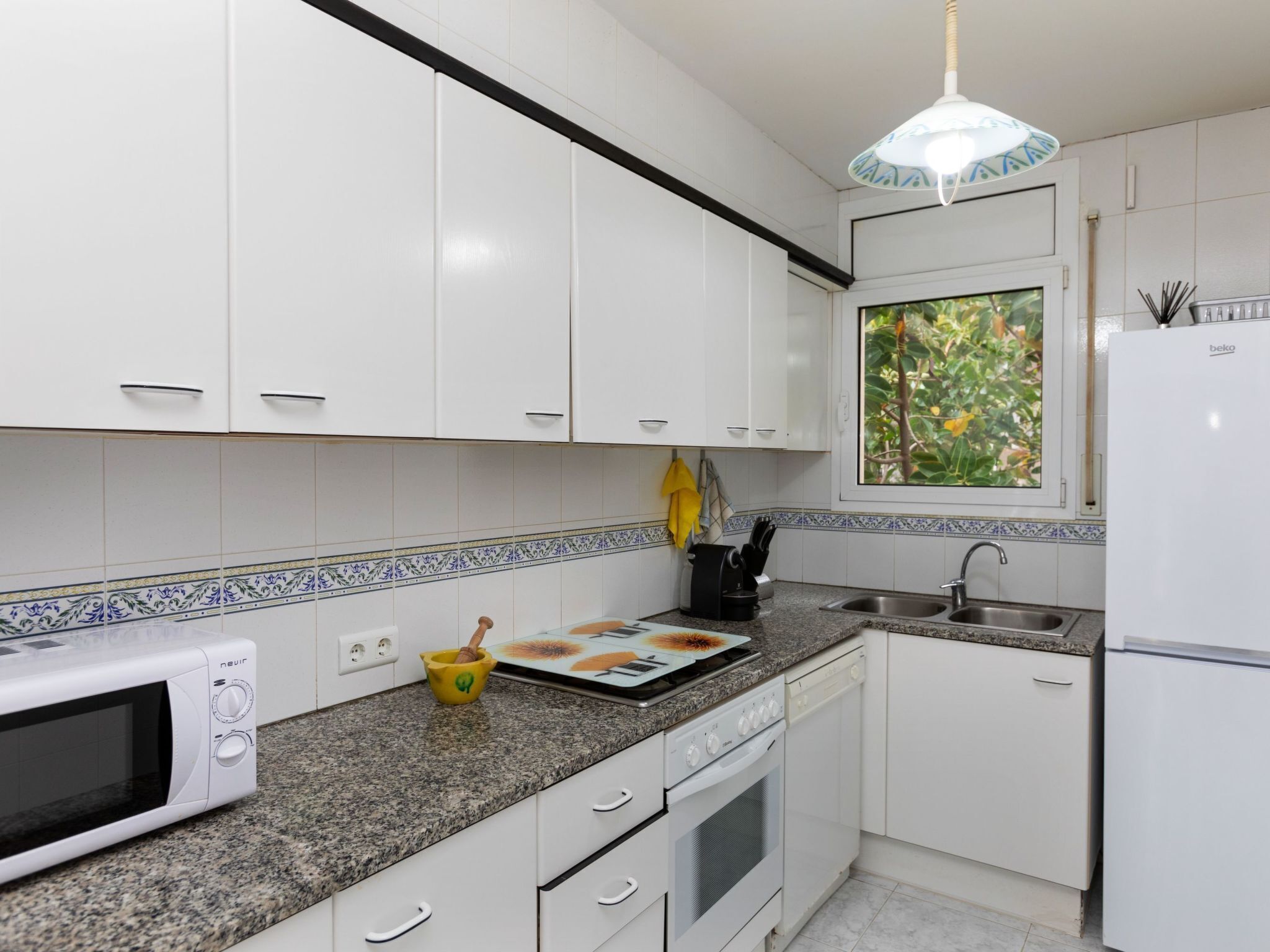 A modern kitchen with white cabinets, a granite countertop, a microwave, a stove, a sink, and a window with a view of greenery.