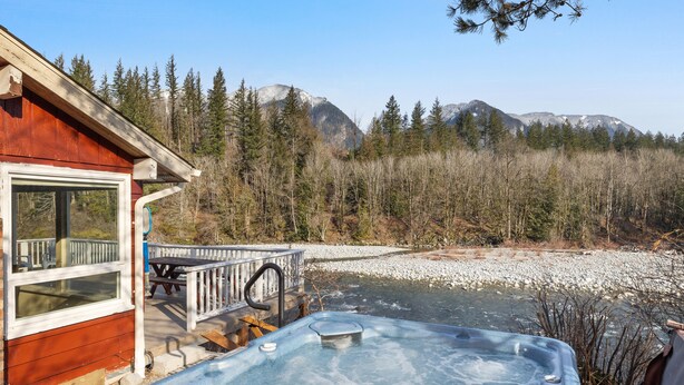 Une cabane en bois dotée d’un bain à remous et d’une terrasse offrant une vue sur une rivière et des montagnes.