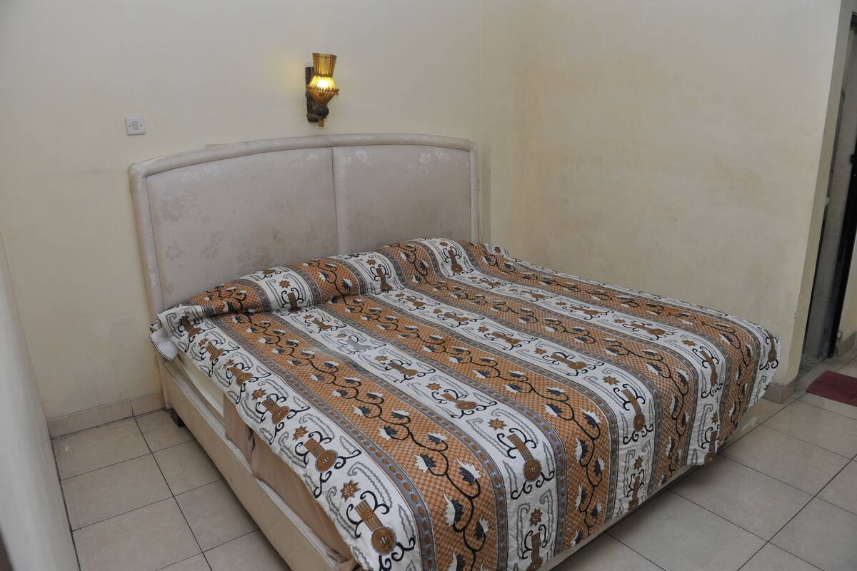 A single bed with a patterned bedspread in a room with a plain wall and a light fixture above the bed.
