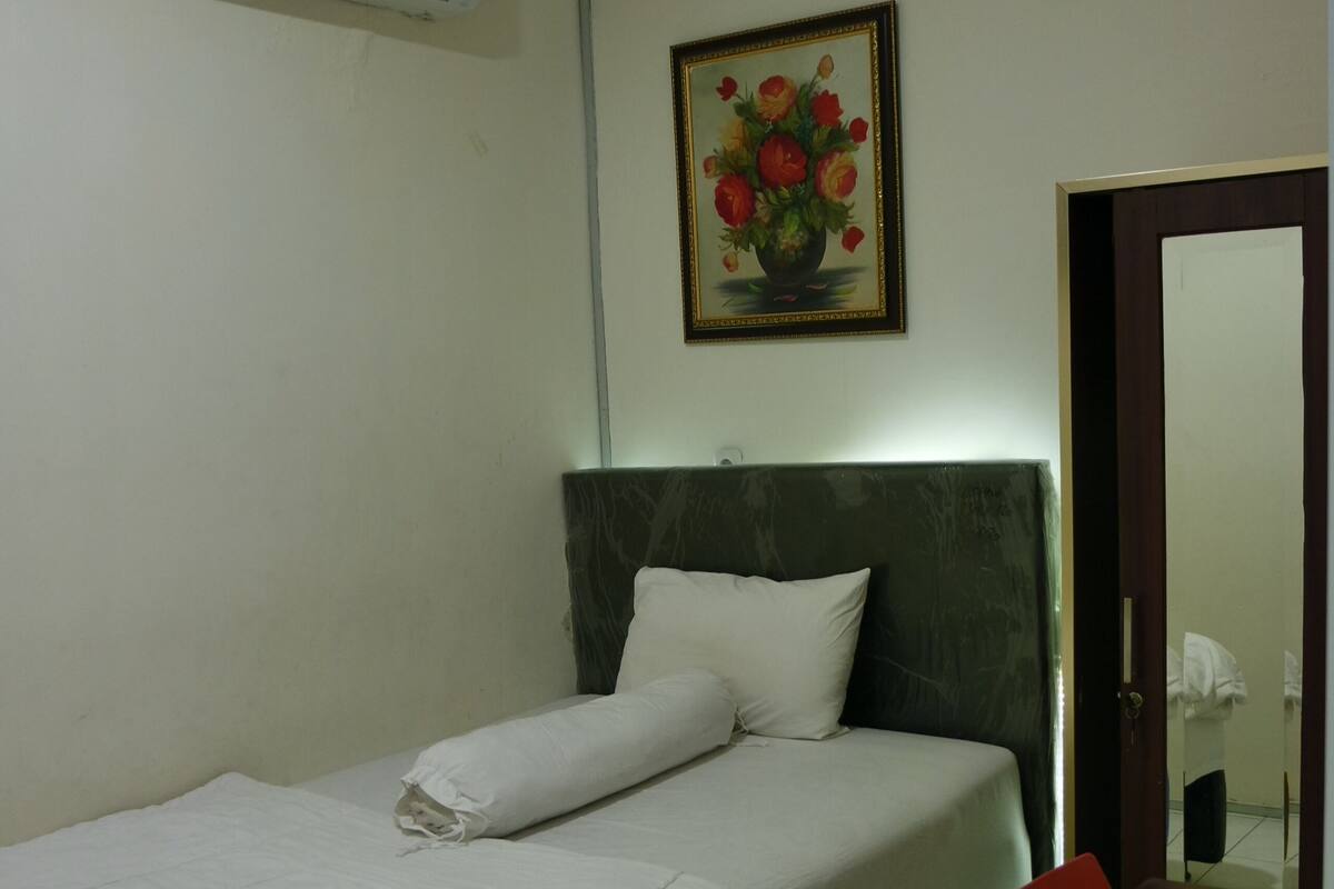 Una cama individual con ropa de cama blanca, una mesita con una silla roja, un aire acondicionado instalado en la pared y un cuadro enmarcado sobre la cama.