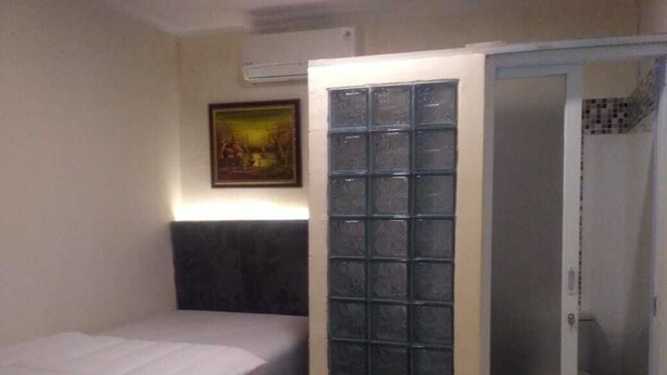 A small, single-bed room with a headboard, a shower area with a glass door, and a framed picture on the wall. - Katalia Tomang (Jakarta)