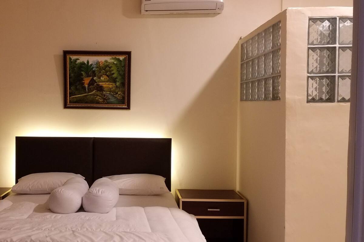 Una cama bien tendida con sábanas y almohadones blancos, un cuadro de un paisaje en la pared y una ventana con diseño de cuadrícula.