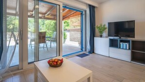 Een moderne woonkamer met een televisie, een witte tafel met een schaal fruit, en een schuifdeur naar een buitenruimte met stoelen.