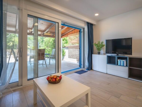 A modern living room with a TV, a white table with a fruit bowl, and a sliding glass door leading to an outdoor area with chairs.