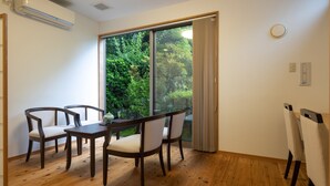 A room with a wooden floor, a dining table with four chairs, a large window with a view of greenery, and an air conditioning unit on the wall. - Temple Hotel Takeibo (Minobu)