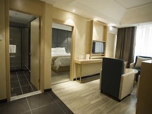 Quarto de hotel com cama, escrivaninha, cadeira, televisão e área de chuveiro.