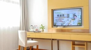 A hotel room with a television on the wall, a desk with a chair, and a window with curtains.