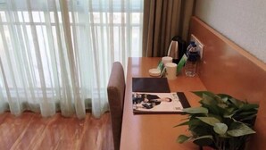 Quarto de hotel com piso de madeira, um sofá, uma mesa com uma revista, uma planta em vaso e uma janela com cortinas translúcidas.