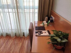 A hotel room with a wooden floor, a sofa, a table with a magazine, a potted plant, and a window with sheer curtains. - Vatica Jieshou City New Passenger Transport Center Hotel (Fuyang)