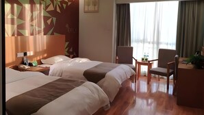 Quarto de hotel com duas camas, uma escrivaninha com cadeira, uma mesinha com cadeira, uma janela com cortinas e um design geométrico na parede.