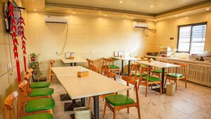 Uma área de refeições com mesas de madeira e cadeiras estofadas em verde, unidades de ar condicionado nas paredes e uma cozinha ao fundo.