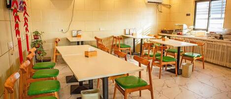 Un comedor con mesas de madera y sillas con cojines verdes, unidades de aire acondicionado en las paredes y una zona de cocina al fondo.