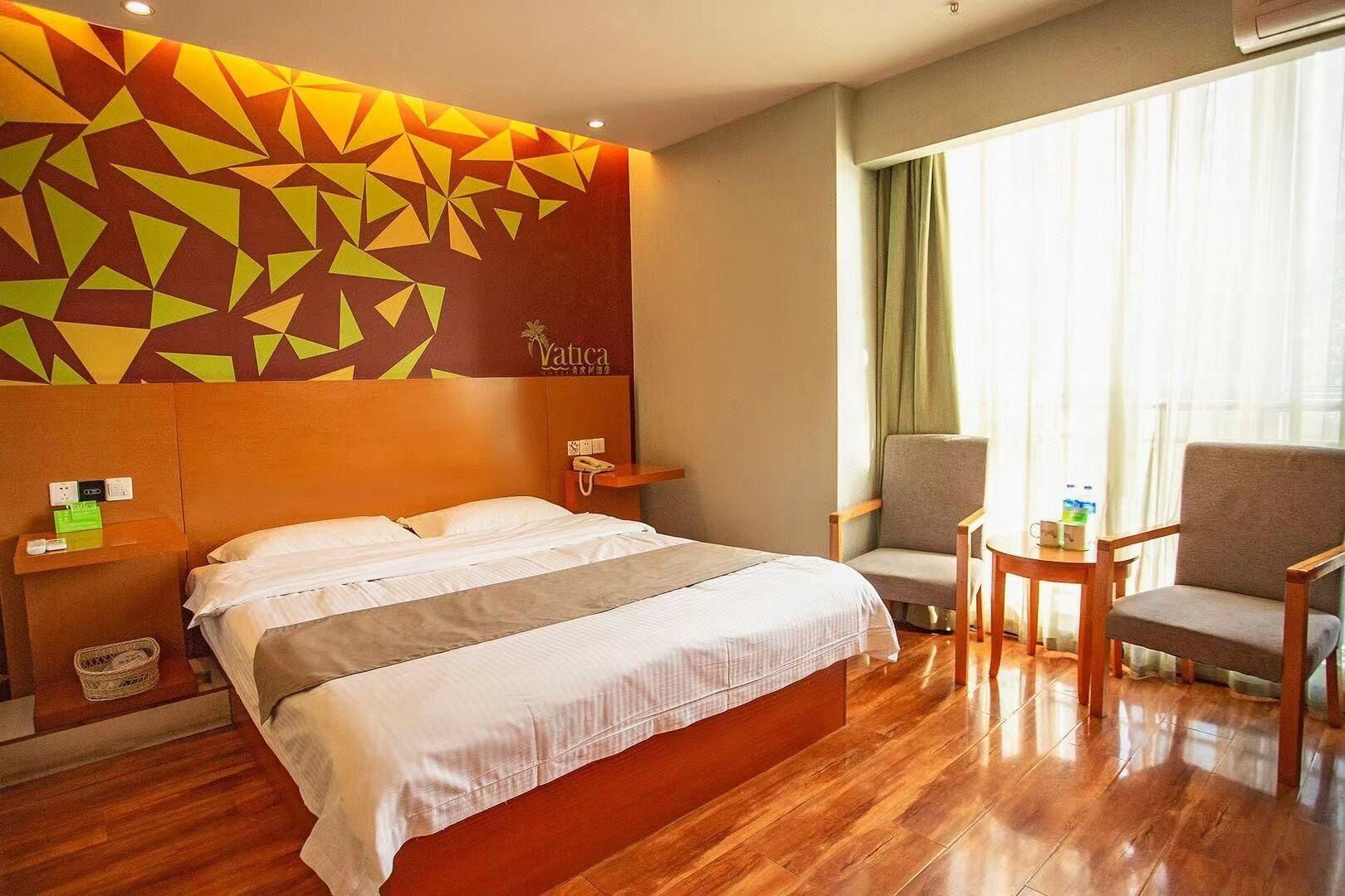A hotel room with a large bed, a headboard with a geometric pattern, a bedside table with a phone, a chair, a small table with a cup, and a window with curtains.