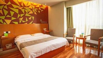 A hotel room with a large bed, a headboard with a geometric pattern, a bedside table with a phone, a chair, a small table with a cup, and a window with curtains.