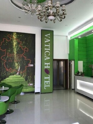 Zona de recepción de un hotel con un mostrador verde, una araña y una pared con un mapa y el nombre del hotel.