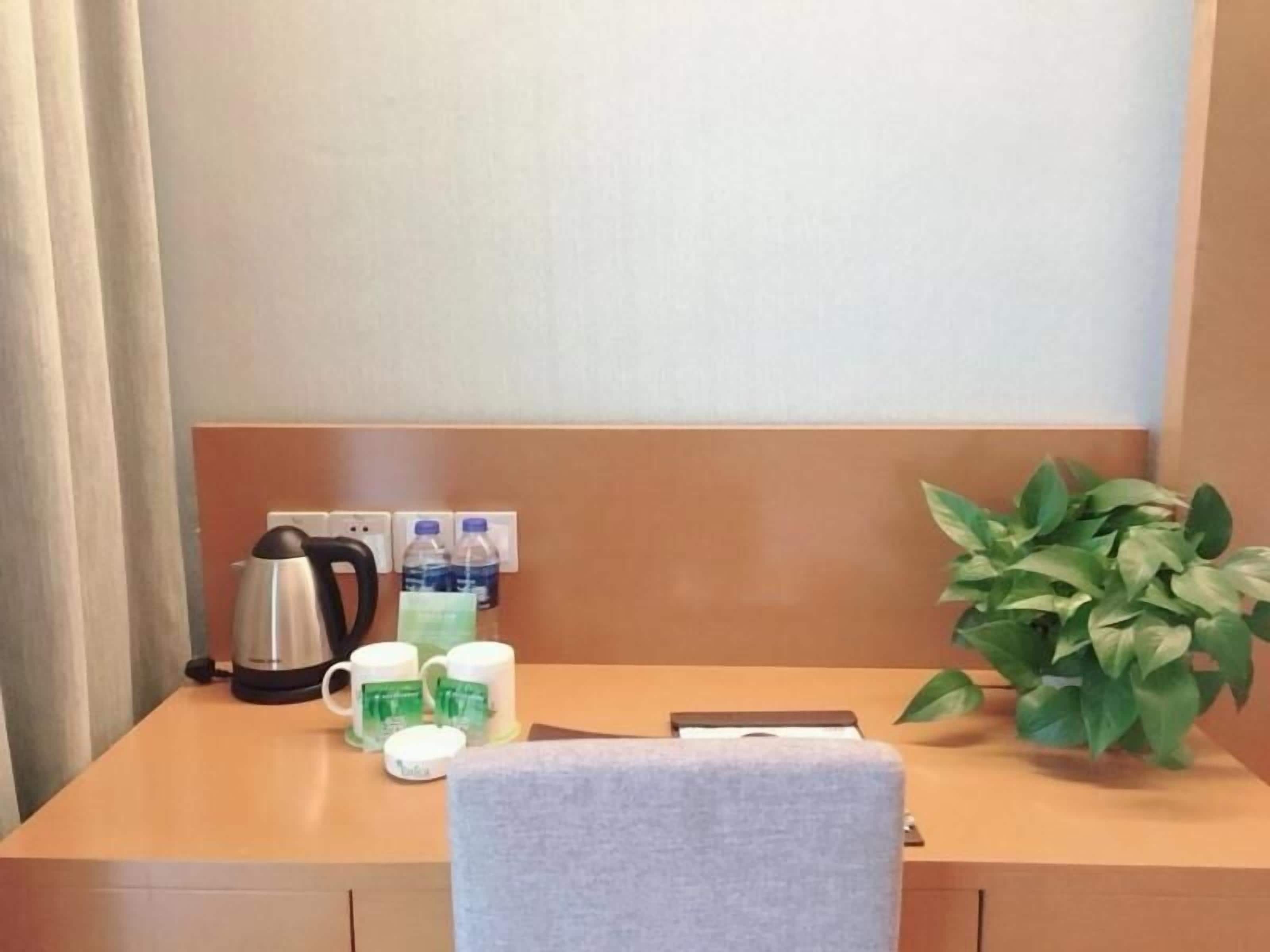 A hotel room with a wooden desk, a kettle, two water bottles, two coffee mugs, a small plant, and a white chair.