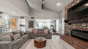 A spacious living room with a gray sectional sofa, a round wooden coffee table, a stone fireplace, and a flat-screen TV mounted on the wall. - 3Kingbeds/Fireplace/Deck/6 Flags/Airport (Grand Prairie)