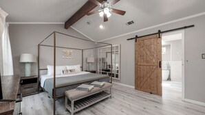 A bedroom with a four-poster bed, a wooden bench, a ceiling fan, and a sliding barn-style door leading to a bathroom. - 3Kingbeds/Fireplace/Deck/6 Flags/Airport (Grand Prairie)