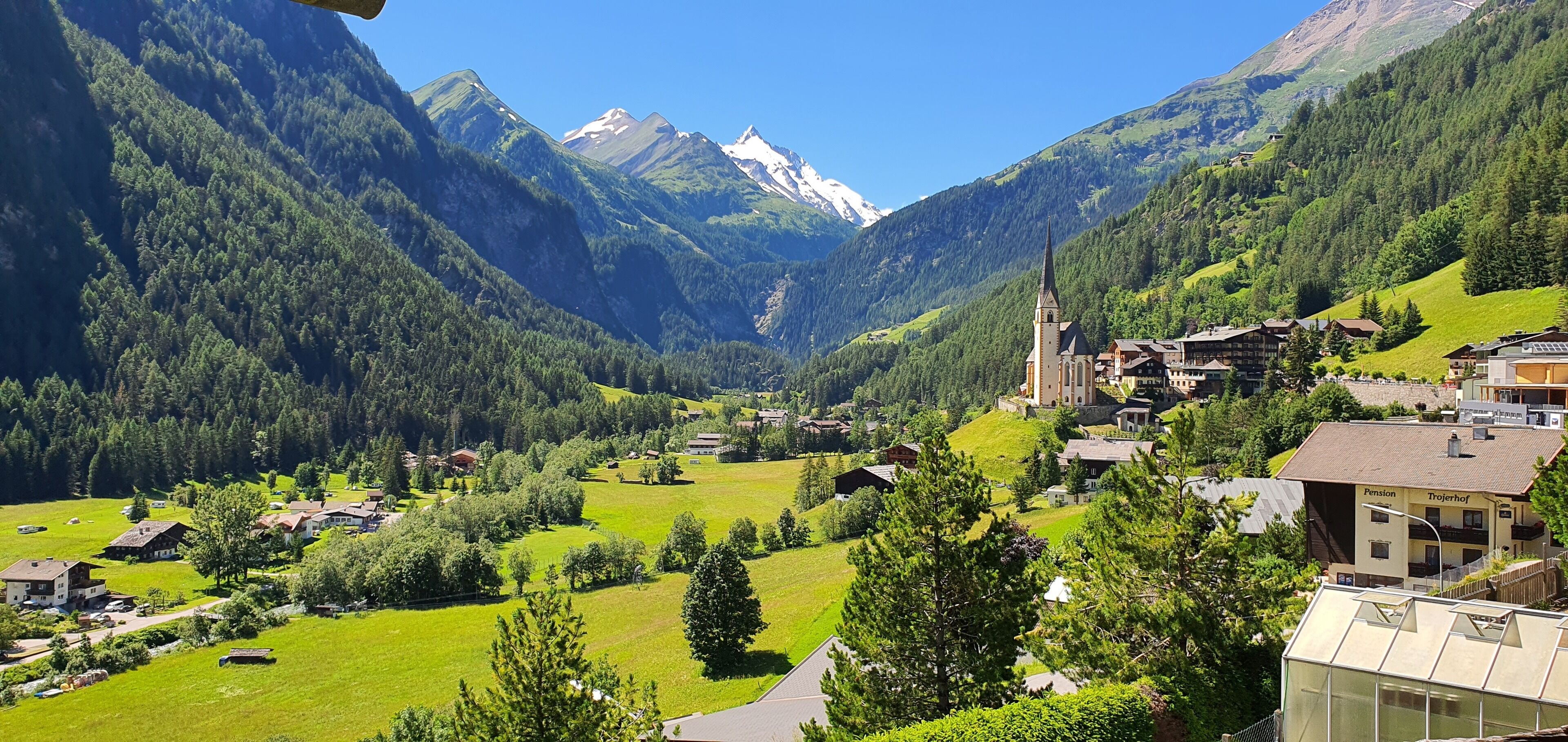 A mountain village with traditional buildings, a church, and a valley with green fields and trees.