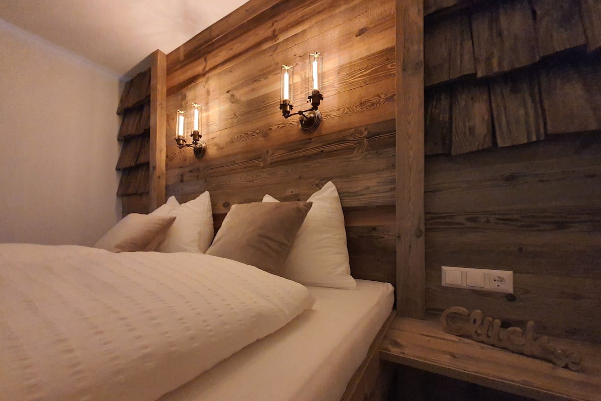 A bed with white bedding and multiple pillows, a wooden headboard with wall-mounted lights, and a visible electrical outlet.
