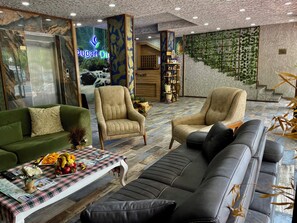 Un vestíbulo moderno con un sofá verde, sillones color beige, una mesa de centro con frutas y un mantel a cuadros.