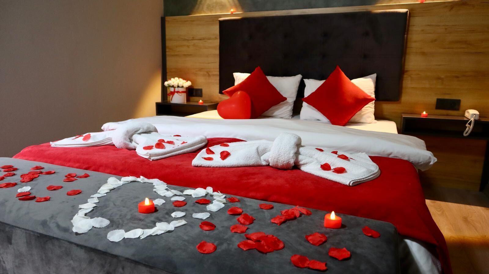 A neatly made bed with red and white pillows, a red and white blanket, and a heart-shaped arrangement of towels and rose petals on a grey bed runner.