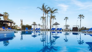 Un espace piscine avec des chaises longues bleues, des palmiers et un ciel d’un bleu éclatant.