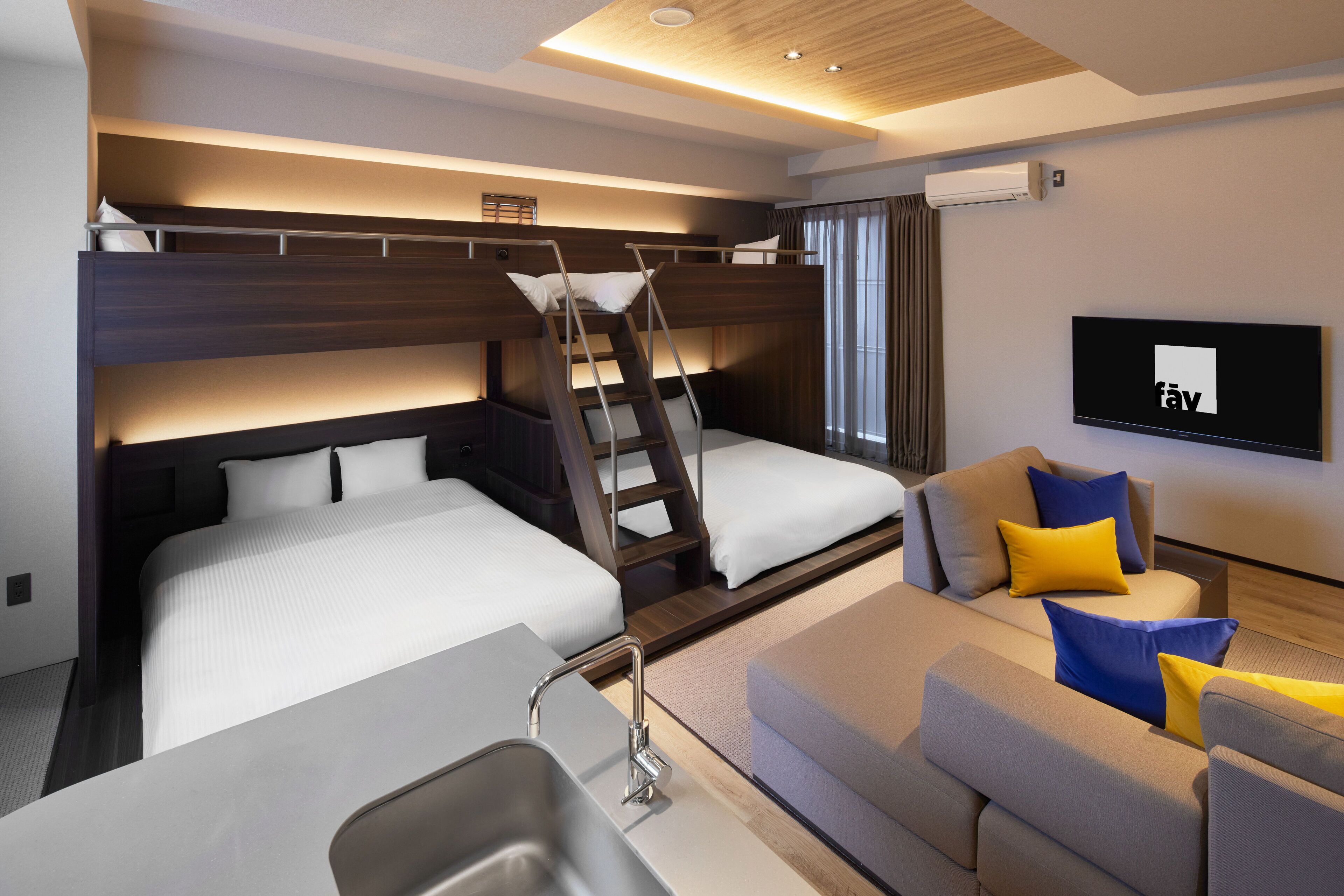 A modern hotel room with bunk beds, a sofa, a TV, and a kitchenette.