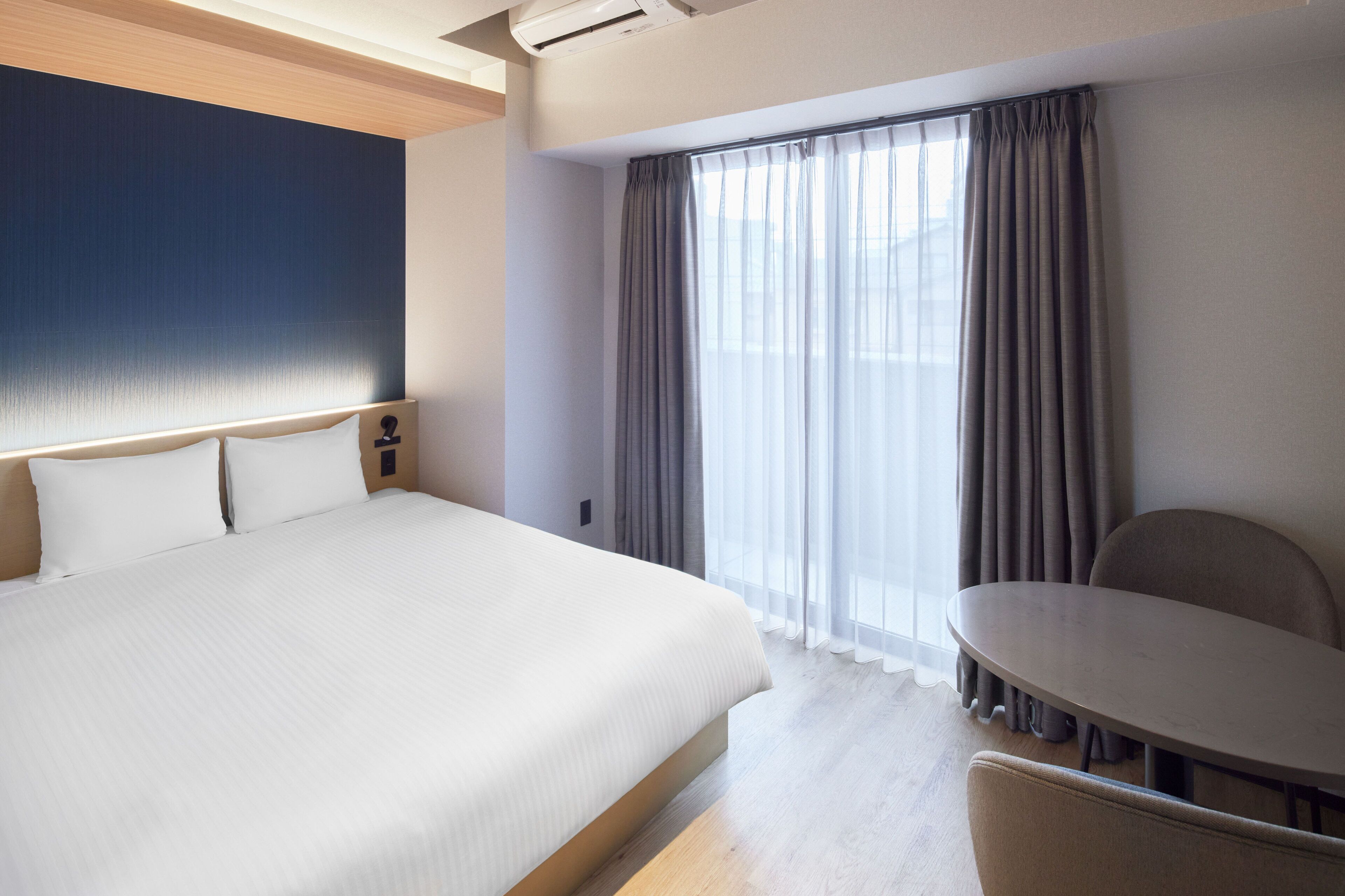 Quarto de hotel com uma cama grande, uma mesa redonda pequena e uma janela com cortinas.