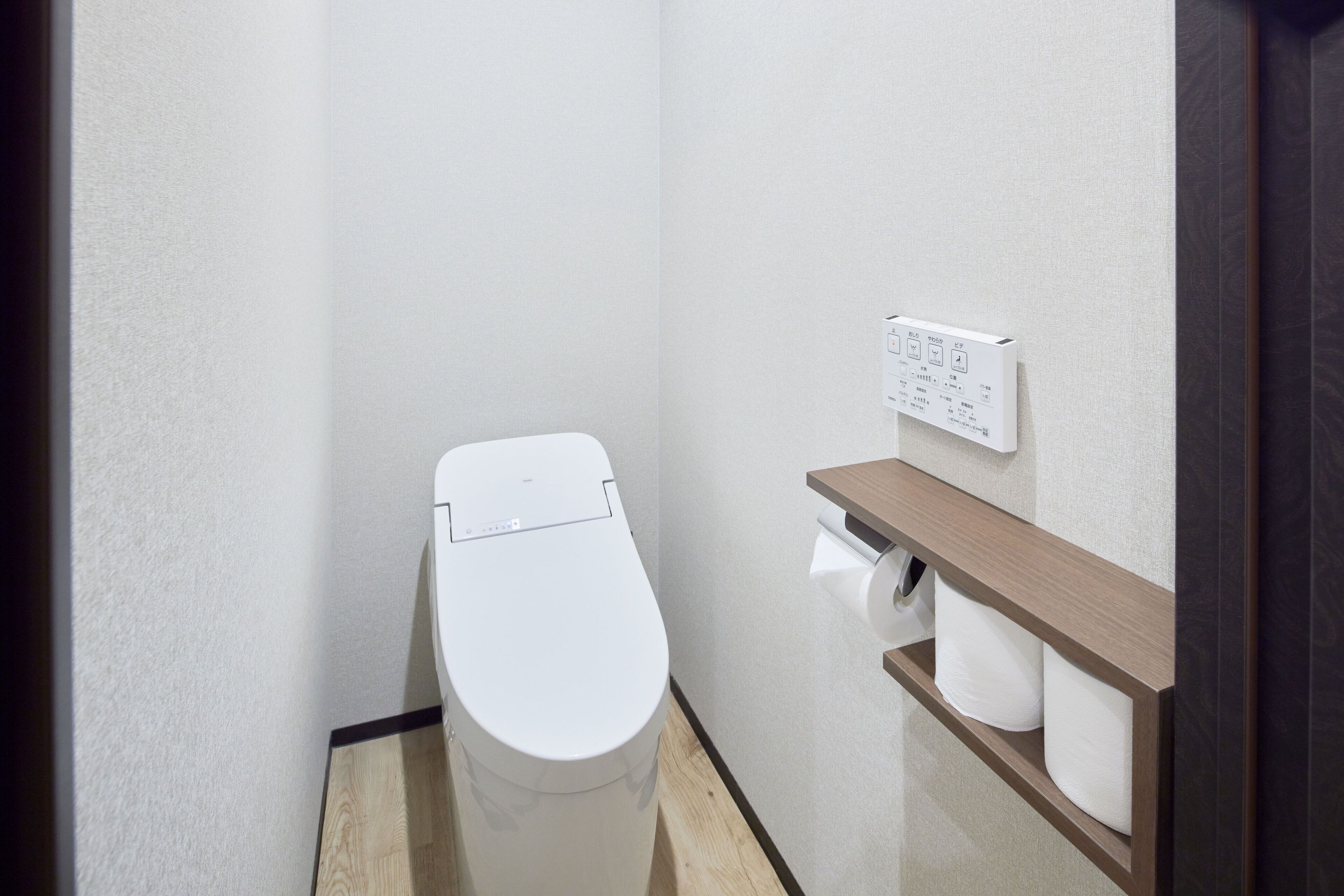A small, modern bathroom with a white toilet, a wooden shelf with toilet paper, and a wall-mounted flush control panel.