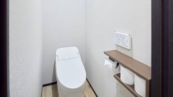 A small, modern bathroom with a white toilet, a wooden shelf with toilet paper, and a wall-mounted flush control panel.