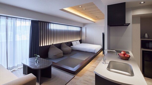 A modern hotel room with a sofa bed, a small table with a vase, a kitchen area with a sink and a bowl of fruit, and a large window with curtains.