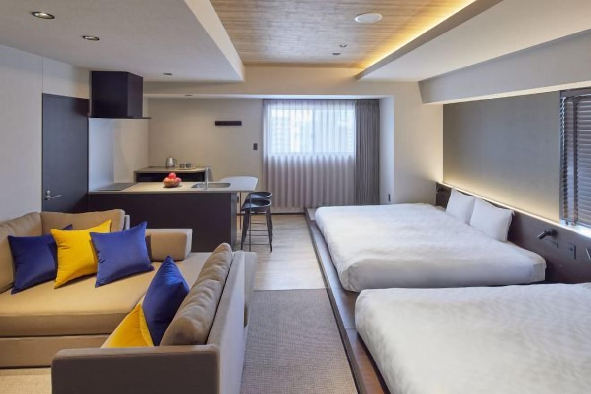Habitación de hotel con sofá, dos camas, kitchenette y un amplio ventanal con cortinas.