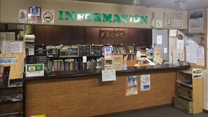 Ein Empfangstresen mit einem Schild, auf dem 'INFORMATION' steht, und einem Schild mit der Aufschrift 'FRONT' darüber. An den Wänden befinden sich verschiedene Aushänge und Schilder, sowie ein Tresen mit Büchern und Ordnern.