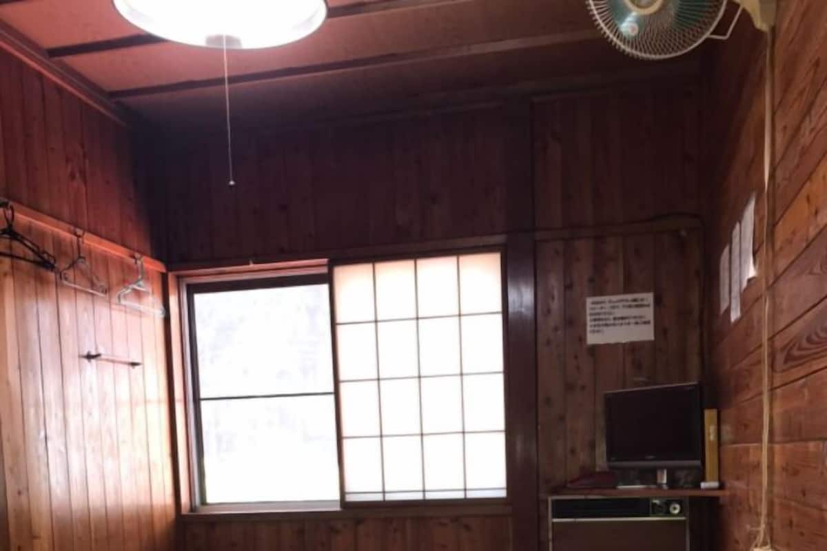 A room with wooden walls, a tatami mat floor, a table, a TV, and a large window with a view.