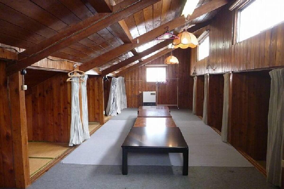 A long, narrow room with wooden walls and a sloped ceiling. There is a central table and a window at the far end.