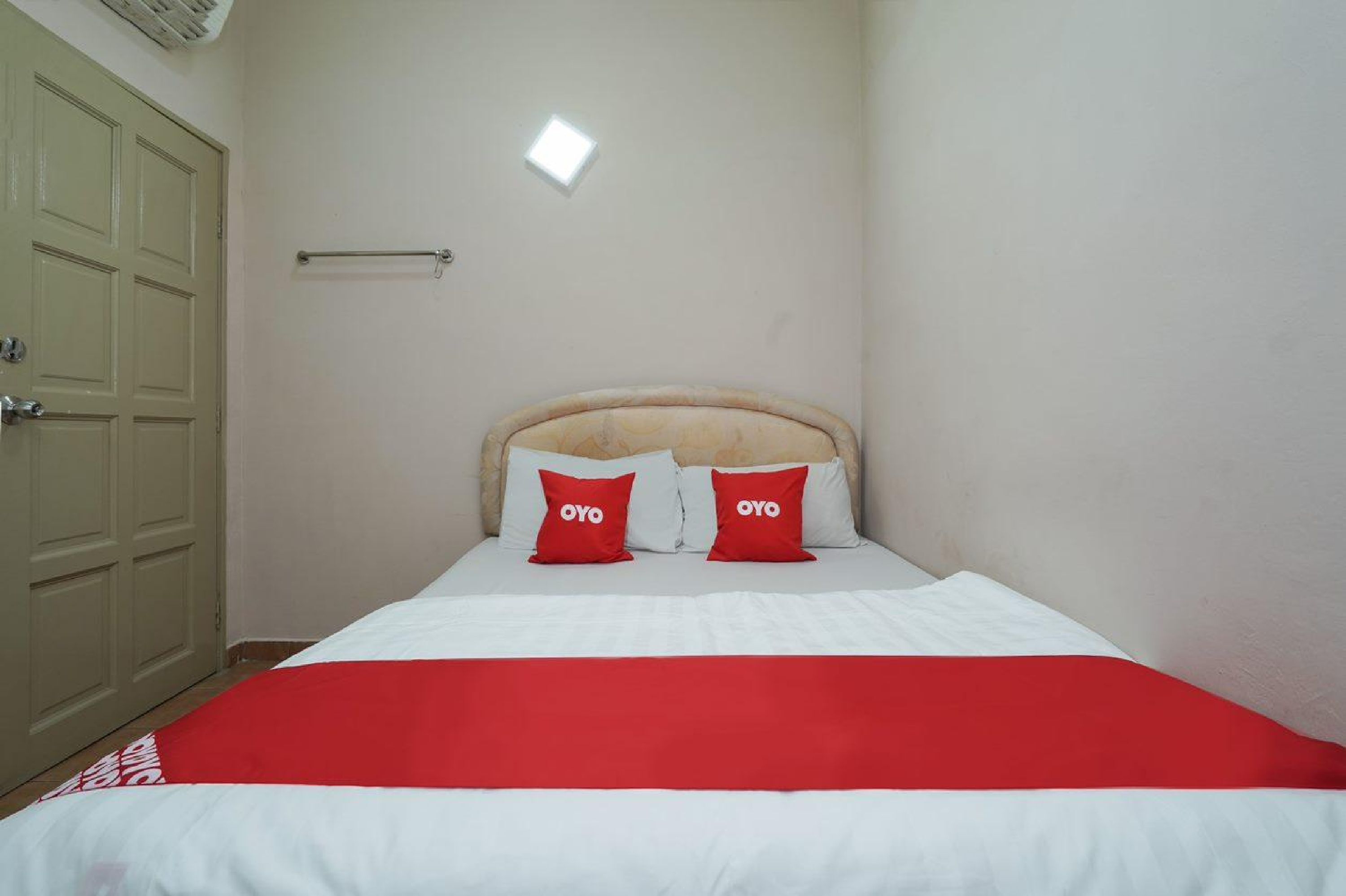 A hotel room with a bed, two red pillows with "OYO" branding, a white bedspread, a beige headboard, a wooden wardrobe, and a small window with a light fixture.