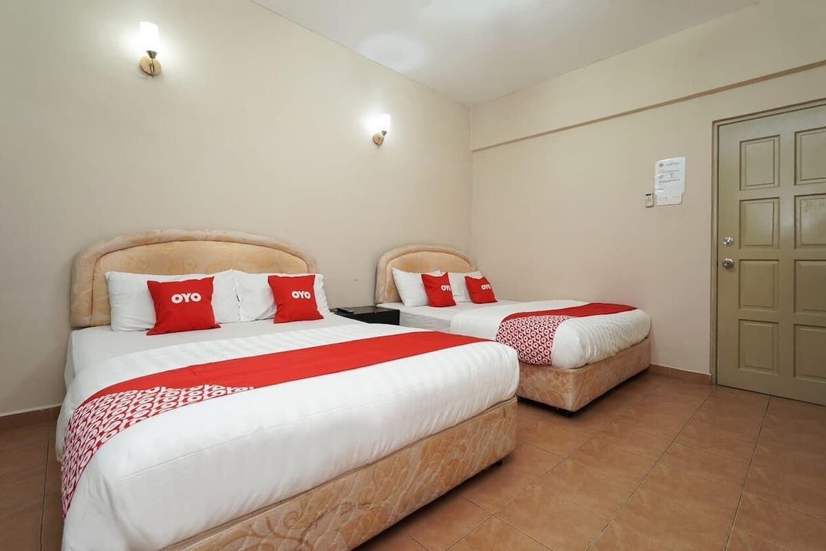 A hotel room with two beds, each with a red and white bedspread and pillows, and a red blanket. There are wall-mounted lights and a door to the right.
