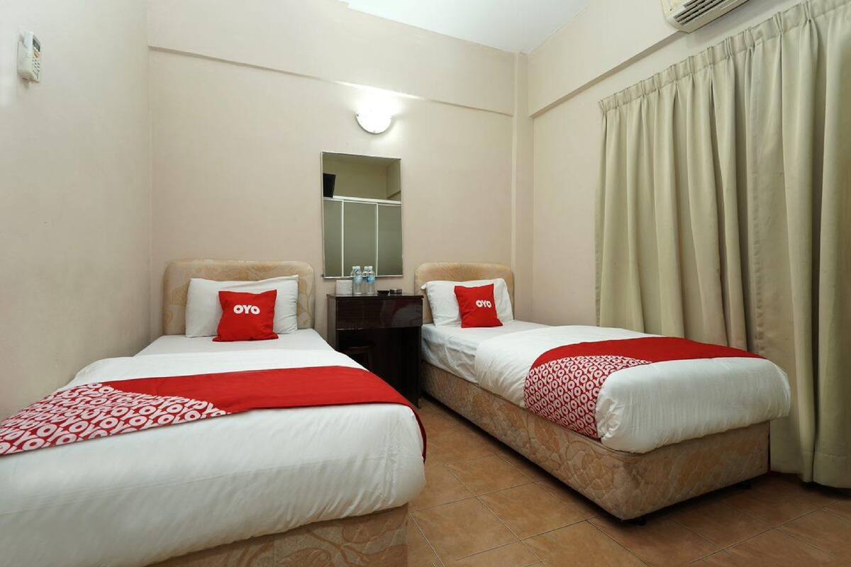 A hotel room with two beds, each with a red pillow and a patterned blanket. There is a small table with a mirror and a lamp between the beds.