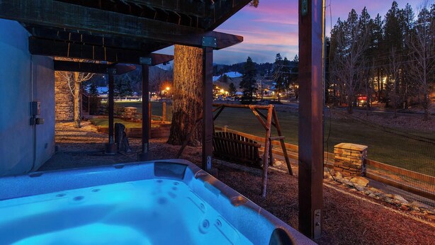 Un jacuzzi con borde de piedra, una terraza de madera y vistas a un parque con árboles y un edificio distante.