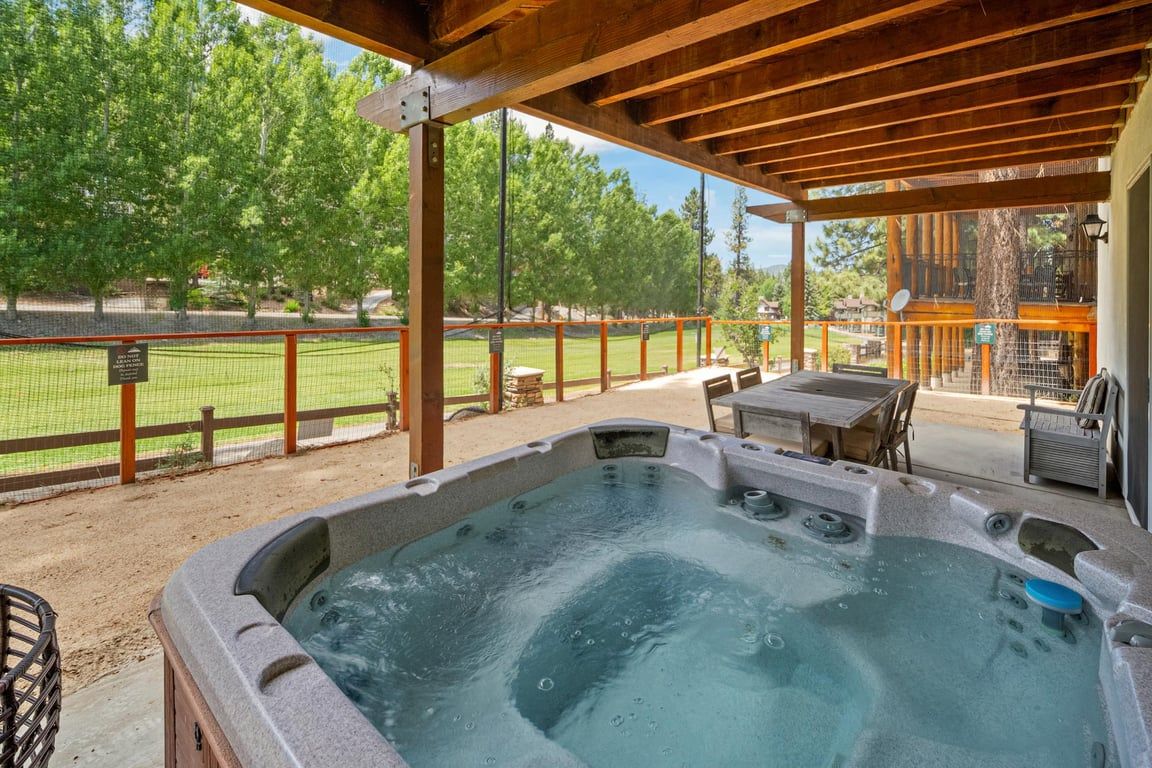 A hot tub area with a wooden pergola, outdoor seating, and a view of a fenced sports field.