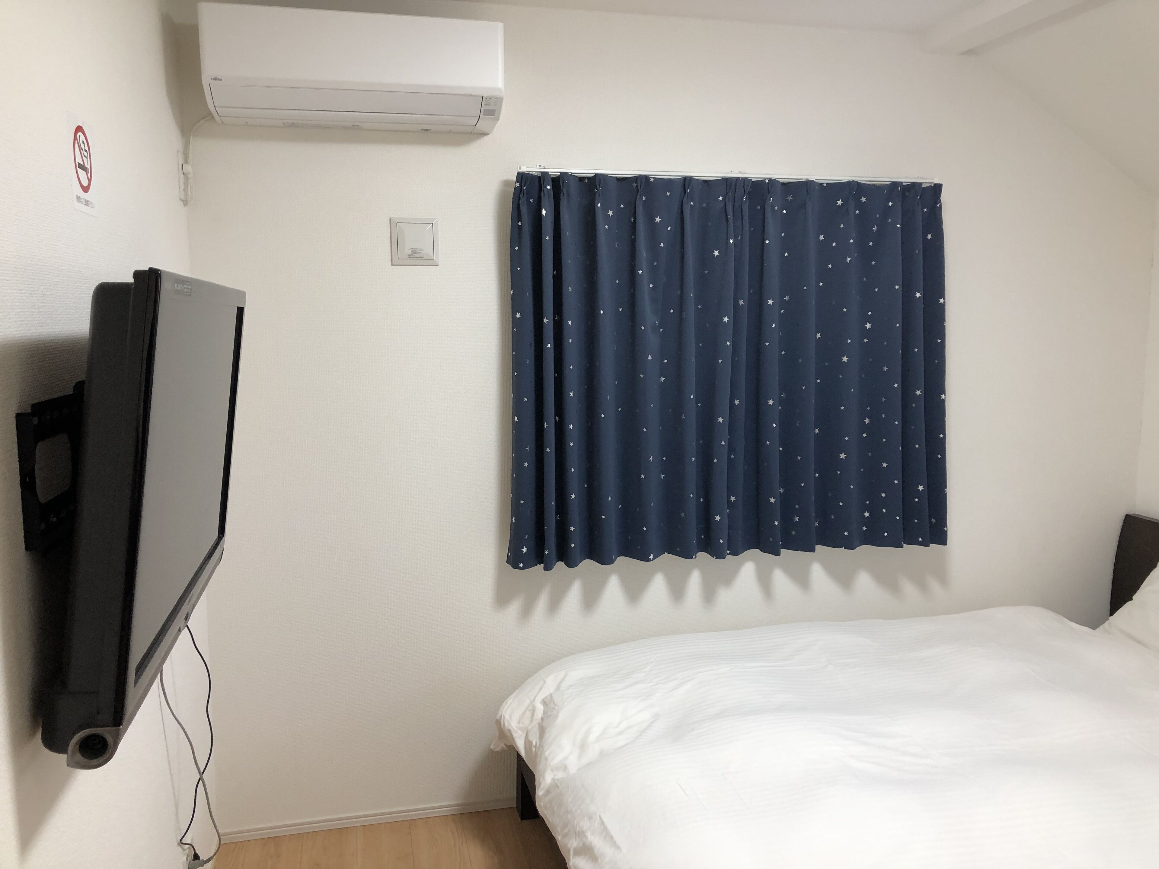 A hotel room with a bed, a TV mounted on the wall, a window with star-patterned curtains, and an air conditioning unit.