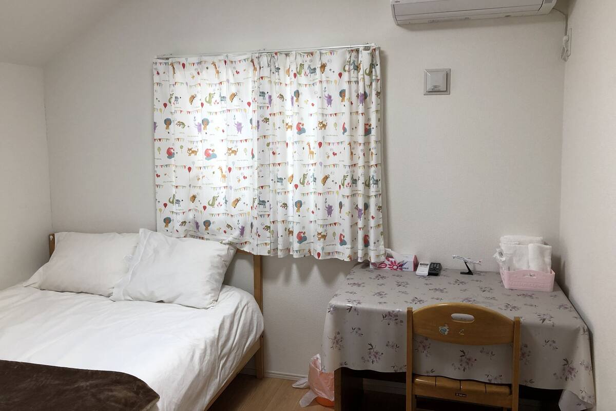 A bedroom with a bed, a table with a floral tablecloth, a chair, and an air conditioning unit on the wall.