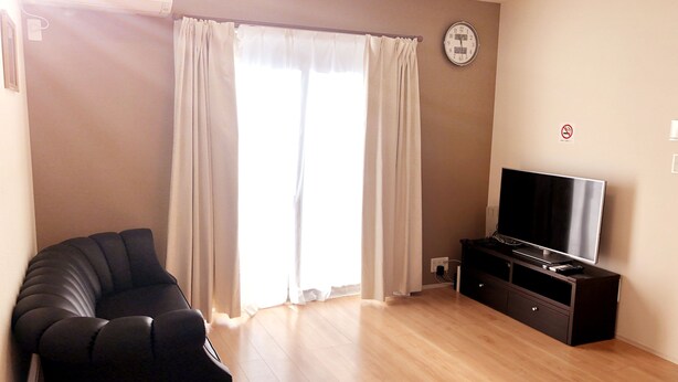 A living room with a black leather sofa, a flat-screen TV on a wooden stand, and a large window with white curtains. - Tokyo Toneri Garden House-Stay (Tokyo)