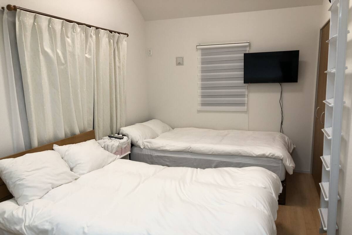 A bedroom with two beds, a TV mounted on the wall, a window with blinds, and a wardrobe with shelves.