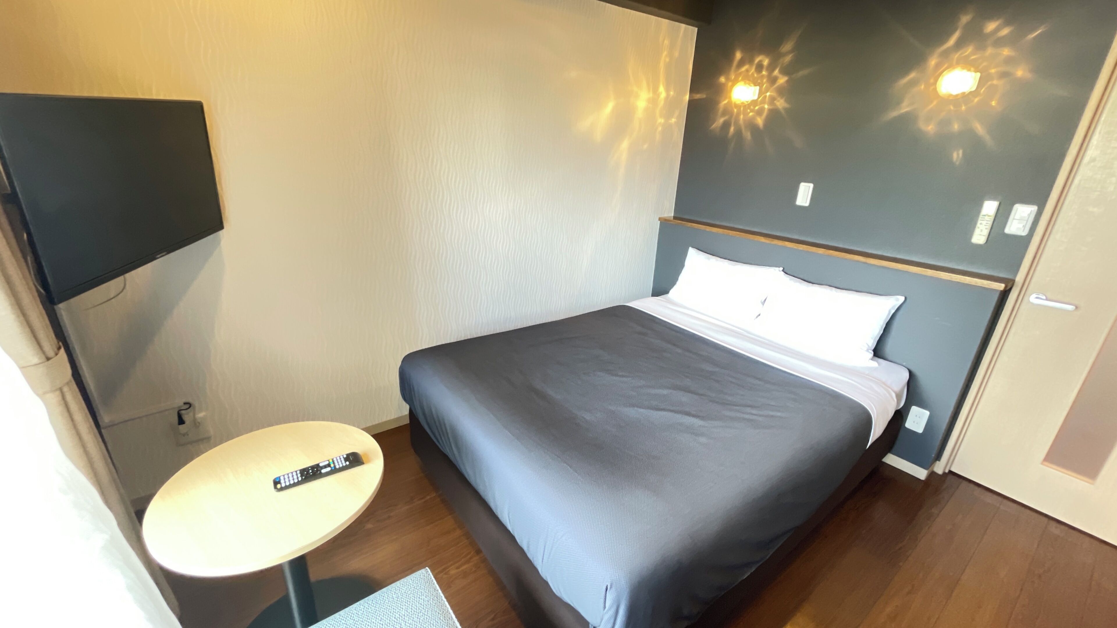A hotel room with a bed, a small round table with a remote control, and a wall-mounted TV.