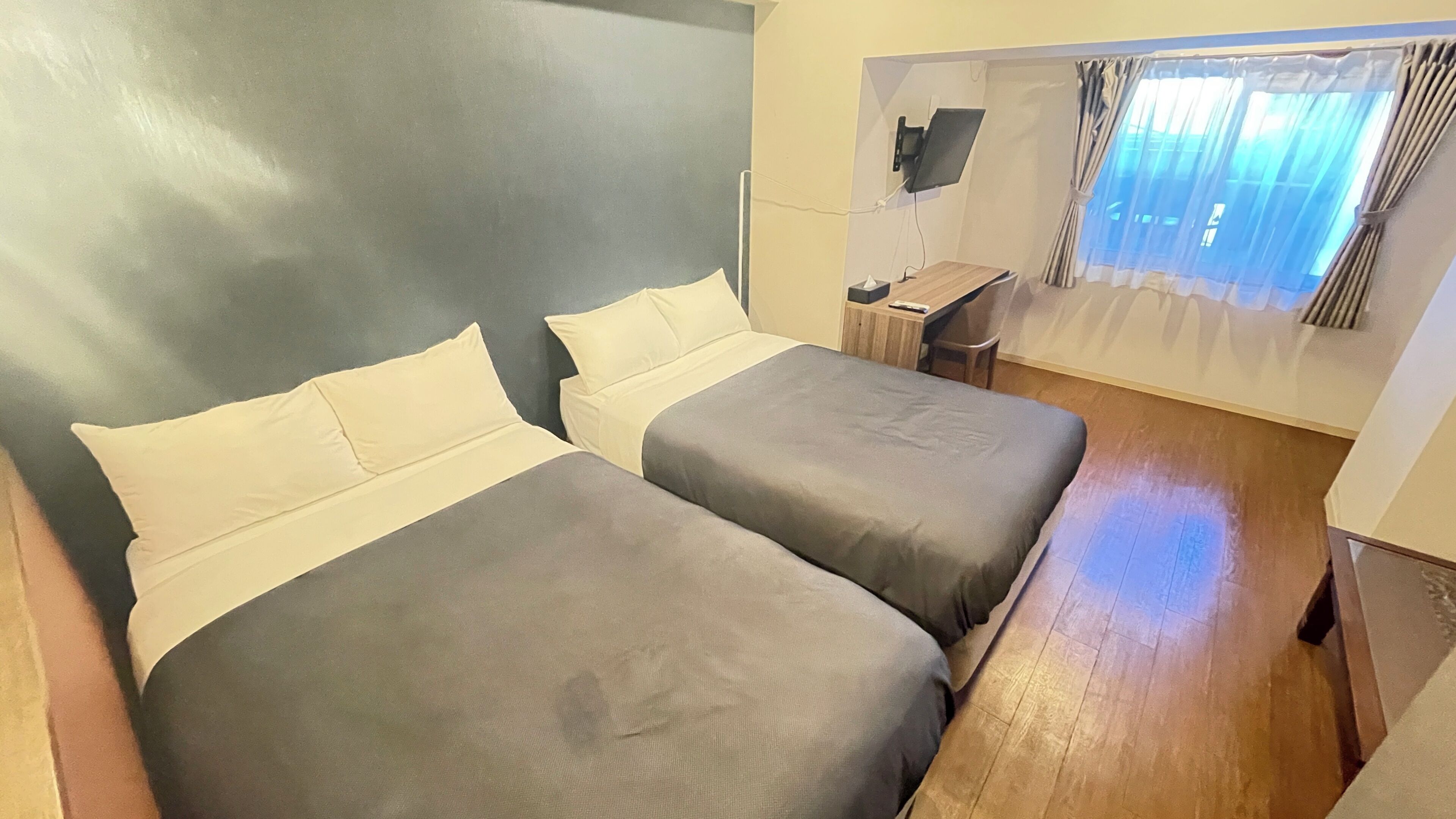 A room with two beds, a desk, a chair, a TV mounted on the wall, and a window with curtains.