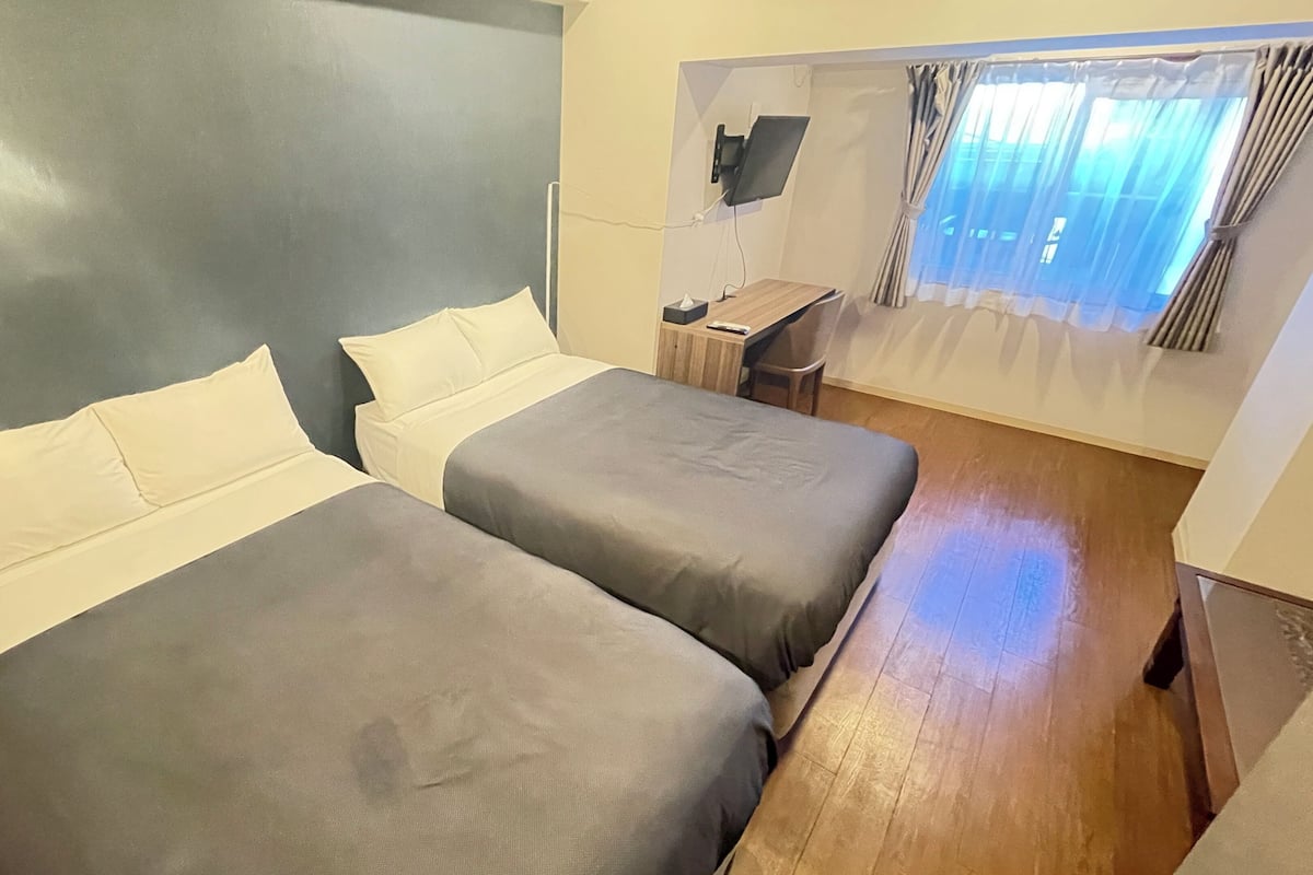 Ein Zimmer mit zwei Betten, einem Schreibtisch, einem Sessel und einem an der Wand montierten Fernseher sowie einem Fenster mit Vorhängen.
