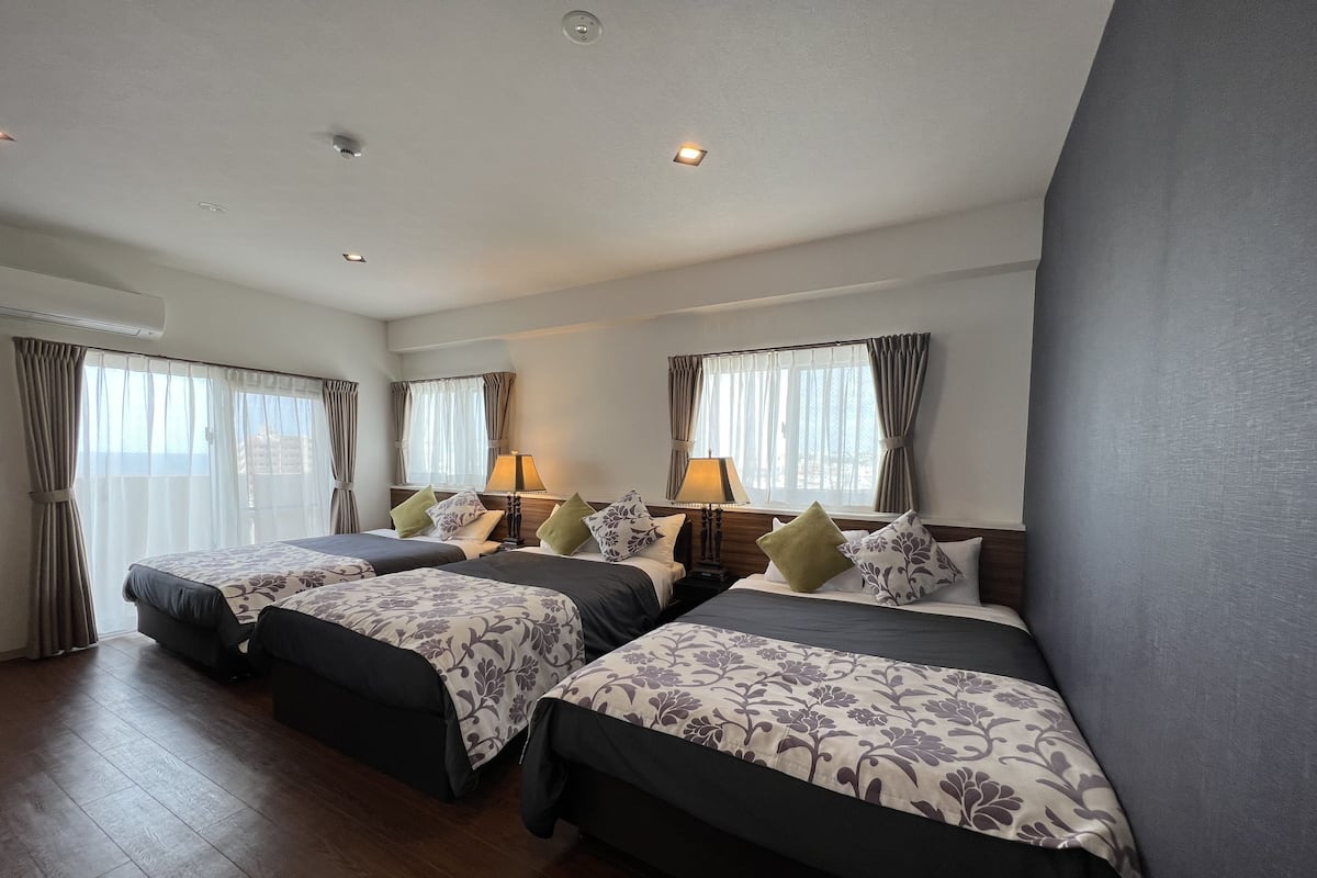 Ein Hotelzimmer mit drei Betten, einem großen Fenster mit Vorhängen, Holzboden und einer Decke mit Einbaustrahlern.